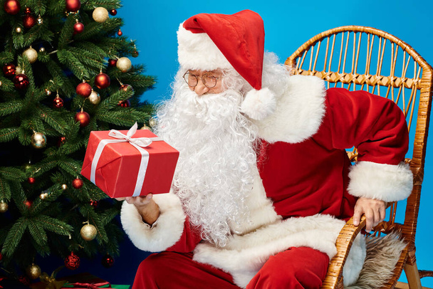 щасливий Санта Клаус з бородою та окулярами сидить у кріслі-качалці з подарунком біля ялинки - Фото, зображення