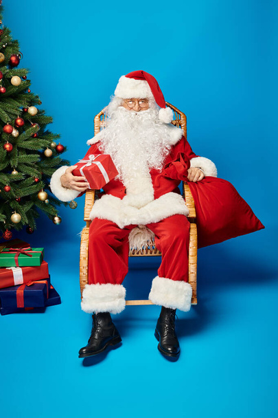 happy Santa Claus in eyeglasses sitting in rocking chair with gift and sack bag near Christmas tree - Photo, Image