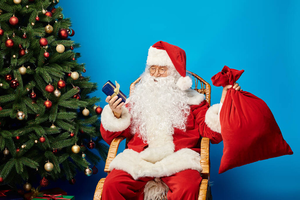 szczęśliwy Święty Mikołaj siedzi w bujanym fotelu z pudełkiem prezentów i worek w pobliżu choinki na niebiesko - Zdjęcie, obraz