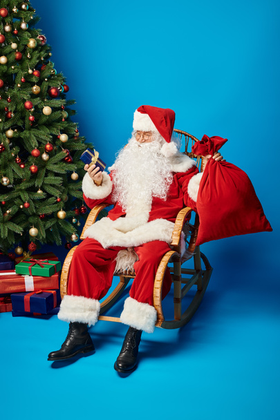glücklicher Weihnachtsmann im Schaukelstuhl sitzend mit Geschenk und Sacksack neben Weihnachtsbaum auf blau - Foto, Bild