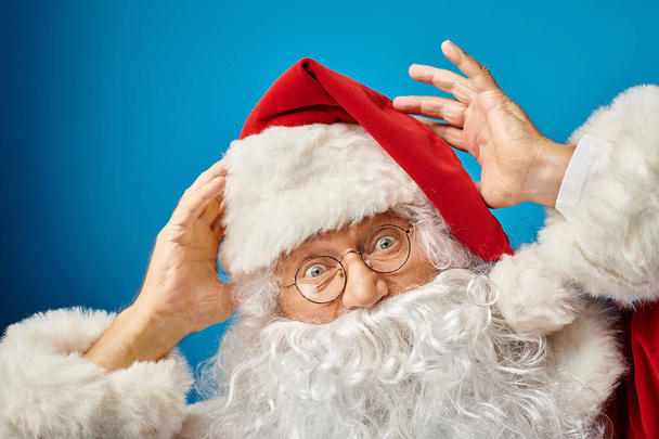 портрет взволнованного Санта-Клауса с белой бородой и очками, смотрящего в камеру на синем фоне - Фото, изображение