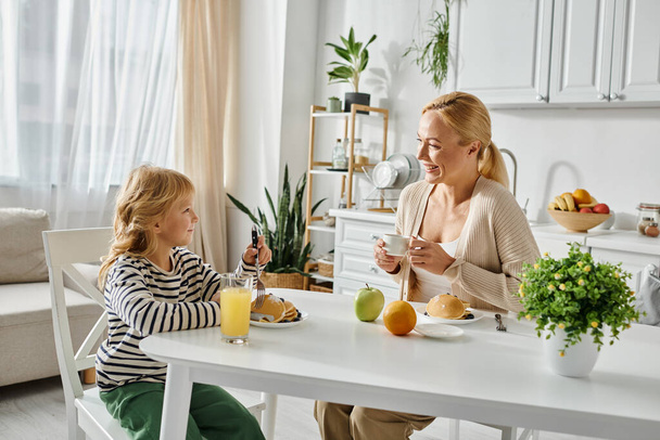 cheerful blonde mother looking at her cute daughter with prosthetic leg having breakfast in kitchen - Photo, Image