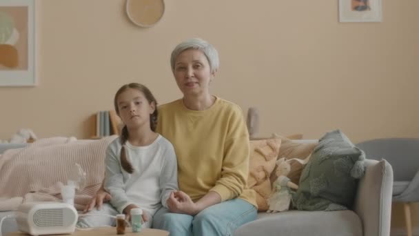 Середній портрет азіатської хворої маленької дівчинки з матір'ю, яка дивиться на камеру, сидячи на дивані у світлій затишній вітальні з інгалятором та ліками на кавовому столі - Кадри, відео