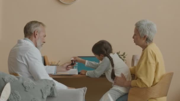 Середній знімок доброго середнього віку кавказького лікаря в лабораторному пальто даючи пляшку таблеток маленькій азіатській дівчинці з мамою, сидячи за столом у світлій вітальні під час домашнього прийому - Кадри, відео