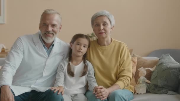 Retrato medio de la niña asiática sentada en el sofá en la sala de estar entre el médico y la abuela, todos sonriendo a la cámara - Imágenes, Vídeo