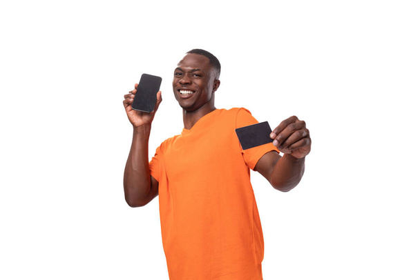 mladý pohledný africký muž oblečený v oranžovém tričku ukazuje kreditní kartu a smartphone na studio bílé pozadí. - Fotografie, Obrázek