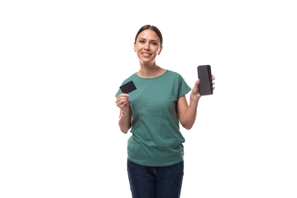 Yeşil, düz kesim tişört giymiş genç, narin bir kadın. Elinde kredi kartı ve akıllı telefon var.. - Fotoğraf, Görsel