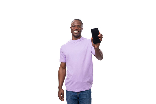 jonge slimme Afrikaanse man gekleed in een lila t-shirt en jeans toont een smartphone mockup. - Foto, afbeelding