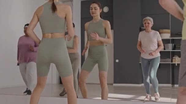 Young female fitness instructor showing exercise in front of mirror to group of senior people during group workout - Footage, Video