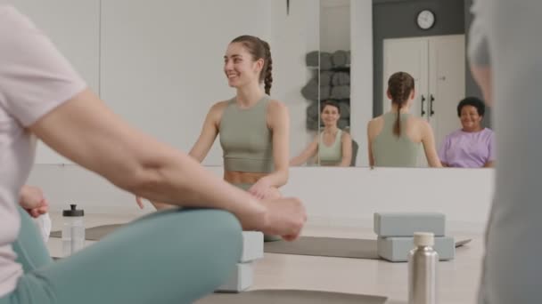 Giovane bella insegnante di yoga femminile seduta con le gambe incrociate sul pavimento in palestra, sorridente e parlando con gruppo di donne prima della pratica - Filmati, video