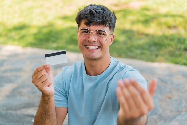 Junger hispanischer Mann, der draußen eine Kreditkarte hält und einlädt, mit der Hand zu kommen. Glücklich, dass du gekommen bist - Foto, Bild