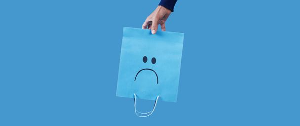 egy férfi kezében egy kék bevásárló papírzacskó, egy szomorú arc festett rajta, fejjel lefelé a kék háttér, panorámás formátumban használni, mint a web banner - Fotó, kép