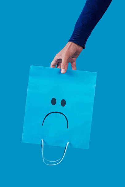 close-up de um homem segurando um saco de papel de compras azul vazio, com um rosto triste pintado nele, de cabeça para baixo em um fundo azul - Foto, Imagem