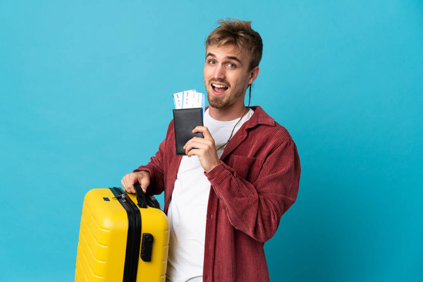 Joven guapo hombre rubio aislado sobre fondo azul en vacaciones con maleta y pasaporte - Foto, Imagen