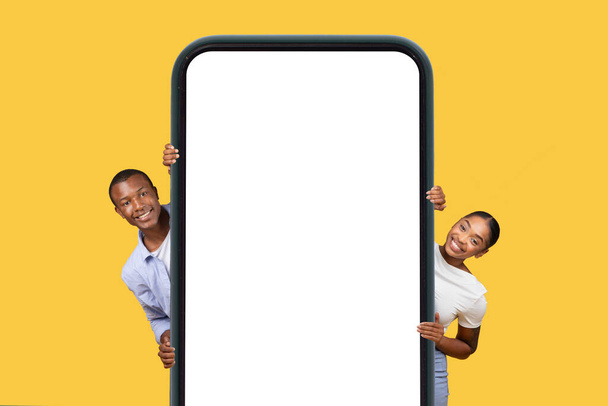 Cheerful black man and woman smiling while playfully peeking out from behind large, vertical blank mobile phone screen over yellow background, mockup - Photo, Image