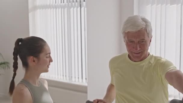 Střední záběr veselý šedovlasý senior muž dělá činky činky s pomocí mladé ženy fitness trenér v tělocvičně - Záběry, video