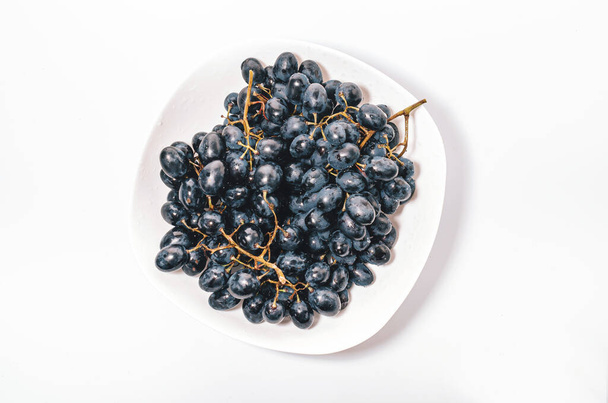 racimos de uvas azules en un plato blanco sobre un fondo blanco. Bayas, cosecha de verano, alimentación saludable - Foto, imagen