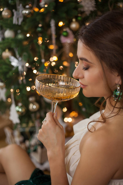 シャンパンのガラスとクリスマスツリーの近くのスタイリッシュな服で美しい若いダークハイレッドの女性. ホリデー。 新年. - 写真・画像