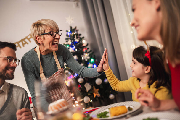 La abuela y su nieta chocan los cinco mientras el resto de la familia disfruta de la cena de Navidad. Familia feliz multi-generación  - Foto, Imagen