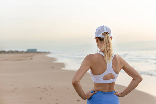 una ragazza magra in calzamaglia blu e un top bianco si staglia sullo sfondo del mare all'alba facendo esercizi, il concetto di uno stile di vita sano. Foto di alta qualità - Foto, immagini