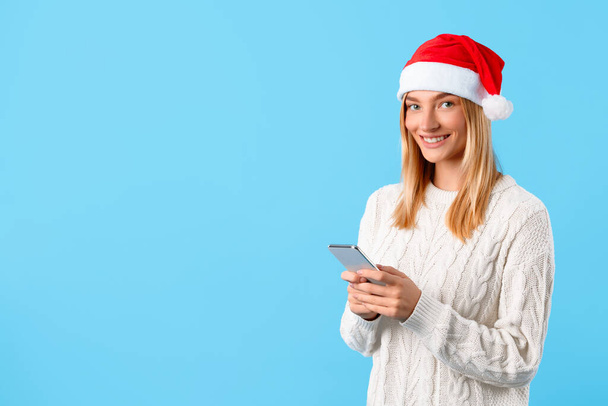Junge Dame mit blonden Haaren mit Weihnachtsmütze lächelt, während sie ihr Smartphone genießt, und verkörpert die festliche Stimmung auf leuchtend blauem Hintergrund, freier Raum - Foto, Bild