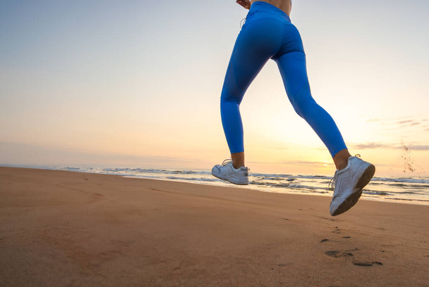 benen van een meisje in blauwe legging en sneakers die bij zonsopgang langs het strand rennen met ruimte voor inscriptie. Hoge kwaliteit foto  - Foto, afbeelding