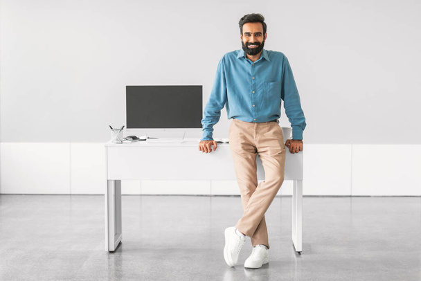 Glimlachend zelfverzekerde Indiase zakenman die aan een modern bureau staat met een blanco computermonitor, wat staat voor professionaliteit en paraatheid. - Foto, afbeelding