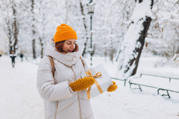 Porträt einer lächelnden stilvollen Frau mittleren Alters mit Geschenk im winterlichen Stadtpark. - Foto, Bild