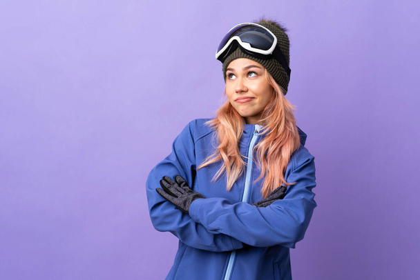 Skier adolescente avec des lunettes de snowboard sur fond violet isolé faire des doutes geste tout en soulevant les épaules - Photo, image