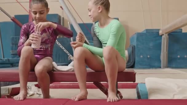 Dwie wielorasowe dziewczynki w trykotach, siedzące na małej równoważni i rozmawiające podczas treningu gimnastyki artystycznej w jasnym, przestronnym studio - Materiał filmowy, wideo