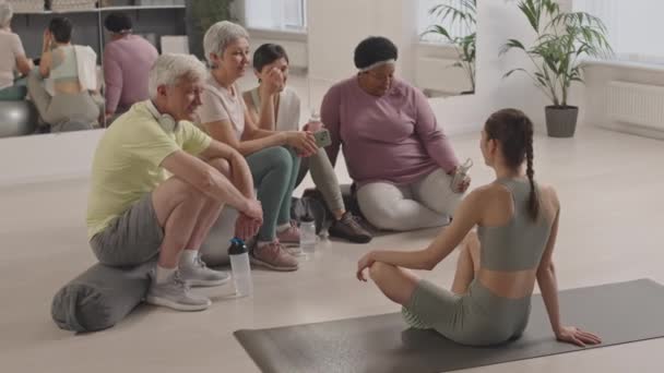 Grupo de personas mayores descansando después del entrenamiento, tomando agua y charlando con una instructora de fitness sentada en una alfombra frente a ellos - Imágenes, Vídeo