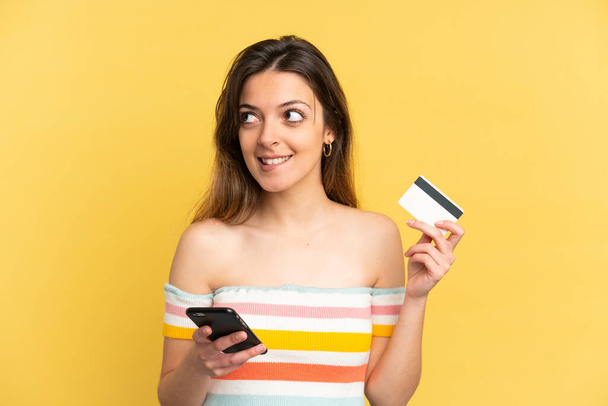 Young caucasian woman isolated on yellow background buying with the mobile with a credit card while thinking - 写真・画像