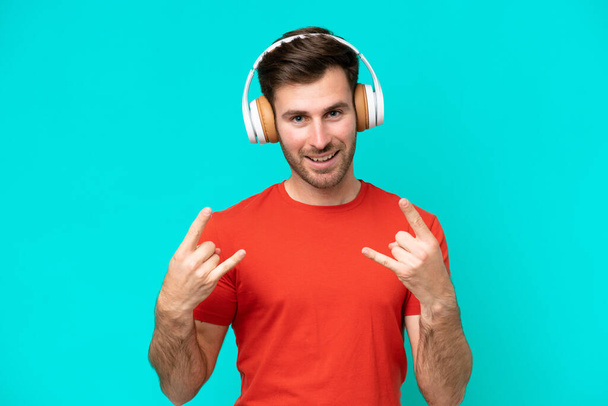 Nuori valkoihoinen mies eristetty sinisellä taustalla kuuntelee musiikkia tehden rock ele - Valokuva, kuva