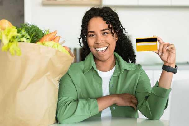 キッチンでオンライン食料品の買い物をお勧めするクレジットカードを示すエキサイティングなミレニアルアフリカ系アメリカ人女性,食料品がいっぱいの紙袋でポーズしながらカメラに微笑みます - 写真・画像