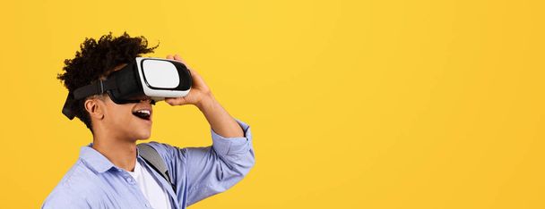 Innoissaan nuori musta mies kihara tukka kokevat virtuaalitodellisuus VR kuulokkeet, hymyillen laajalti katsot vapaata tilaa kirkkaan keltainen tausta, panoraama - Valokuva, kuva