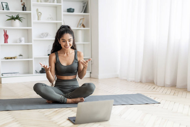 Молодая индийская женщина инструктор yoga тренера yoga имеет online класс с студентом, спортивная женщина сидя на циновке на дому, смотря на экран лэптопа, улыбаясь и жестикулируя, место для копирования. Онлайн тренировки - Фото, изображение