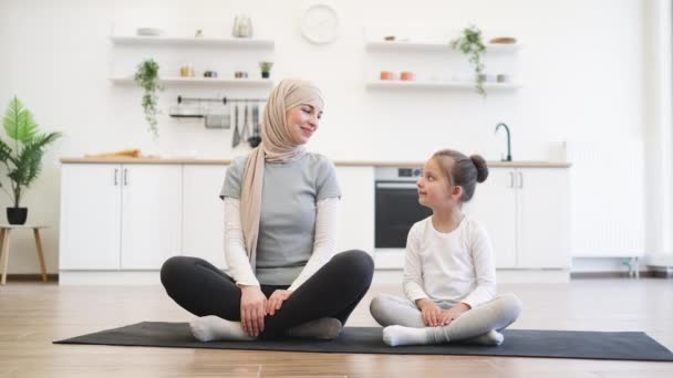 Muslim female with little daughter in sports outfit and closed eyes holding folded palms overhead while meditating in crossed-legged position in fitness studio. Healthy family practicing yoga together - Záběry, video
