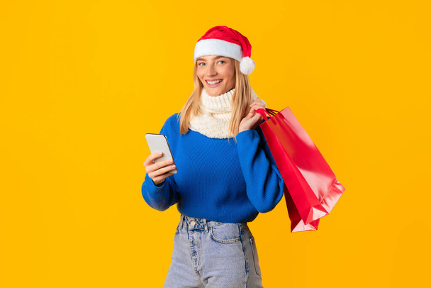 Lächelnde Frau mit Weihnachtsmütze mit Smartphone in der Hand, rote Einkaufstüten in der Hand, fröhlich posierend vor leuchtend gelbem Hintergrund, verkörpert Urlaubsstimmung - Foto, Bild