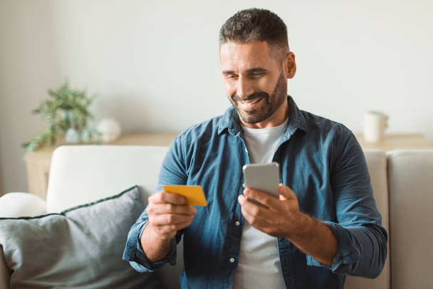 Улыбающийся покупатель средних лет использует свой телефон, чтобы совершать покупки в Интернете, держит кредитную карту в руке, сидя в повседневной домашней обстановке на диване. Концепция цифрового потребления и электронной коммерции - Фото, изображение