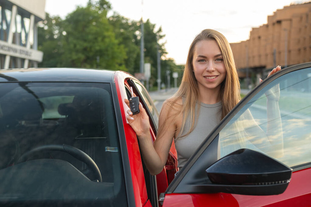 Donna con lunghi capelli biondi in piedi con chiave vicino alla nuova auto rossa in strada. - Foto, immagini