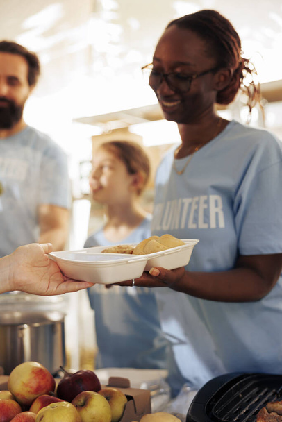 Detailed picture shows volunteers providing free food to the hungry, homeless, and refugees. Close up of female voluntary person distributing meals, providing assistance and sharing non-perishables. - Photo, Image