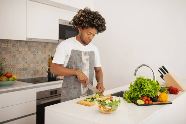 Молодой черный человек в фартуке готовит здоровый вегетарианский ужин, режет овощи для салата, воплощает образ жизни оздоровления и потери веса, стоя в современном помещении кухни - Фото, изображение