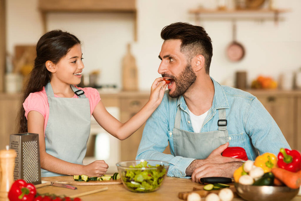 En un ambiente cálido en el hogar, una joven alegre le da rodaja de tomate fresco a su padre encantado durante la sesión de cocina saludable divertida e interactiva - Foto, imagen