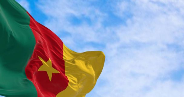 Η εθνική σημαία του Καμερούν με ζωηρά πράσινα, κόκκινα και κίτρινα χρώματα και ένα κεντρικό κίτρινο αστέρι κυματίζει μεγαλοπρεπώς στον άνεμο σε ένα φόντο ενός καθαρού γαλάζιου ουρανού - Φωτογραφία, εικόνα