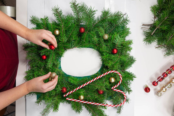 Κορυφαία προβολή γυναικεία χέρια που φτιάχνουν ένα γιορτινό στεφάνι, υφαίνοντας χριστουγεννιάτικες μπάλες. Κόκκινο-και-λευκό καραμέλα μπαστούνια σε πράσινα κλαδιά έλατο με στολίδια Χριστούγεννα.DIY Χριστουγεννιάτικα στεφάνια - Φωτογραφία, εικόνα