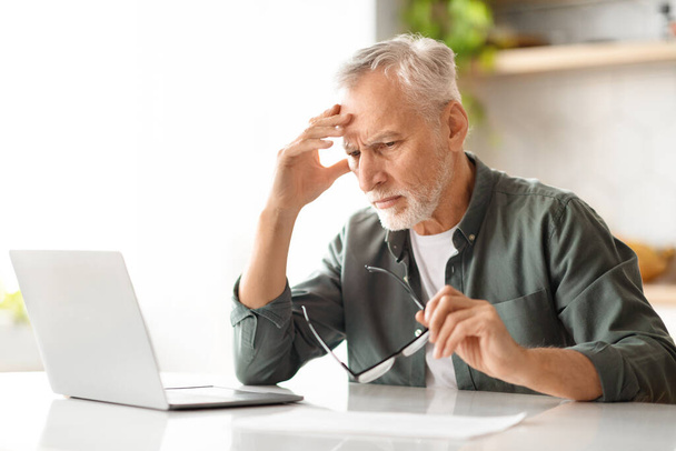 Portrait of stressed senior man using laptop while sitting at desk in kitchen interior, upset elderly gentleman suffering problems with computer, having troubles with freelancing on retirement - Photo, Image