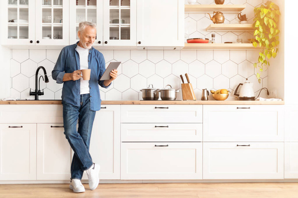 Porträt eines lächelnden älteren Mannes mit digitalem Tablet und Kaffee, der sich in der Küche entspannt, ein glücklicher älterer Mann, der auf modernen Geräten im Internet surft und Heißgetränk genießt, Online-Nachrichten liest, Kopierraum - Foto, Bild