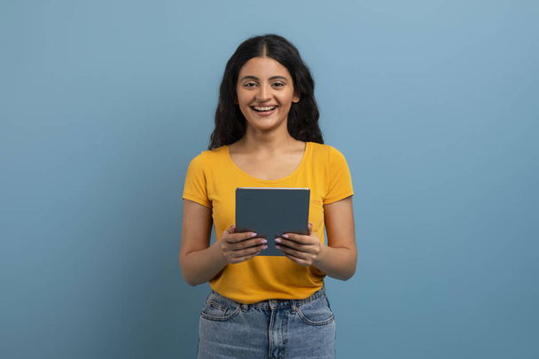 Retrato de la joven dama india alegre usando tableta digital para mensajería con amigos, mensajes de texto de estudiante mujer feliz en la almohadilla mientras está de pie sobre fondo de estudio azul, aprendizaje en línea, espacio de copia - Foto, imagen