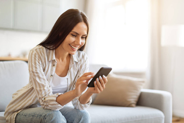 Uśmiechnięta młoda kobieta korzystająca ze smartfona podczas odpoczynku na kanapie w domu, Happy Millennial Kobieta siedząca na kanapie i korzystająca z telefonu komórkowego, zakupy online lub przeglądanie sieci społecznych, Kopiuj przestrzeń - Zdjęcie, obraz