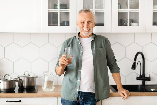 Улыбающийся пожилой человек со стеклом воды в руке позирует в интерьере кухни дома, счастливый красивый пожилой джентльмен наслаждаясь здоровым напитком, глядя на камеру, оздоровительный концепт, копировать пространство - Фото, изображение